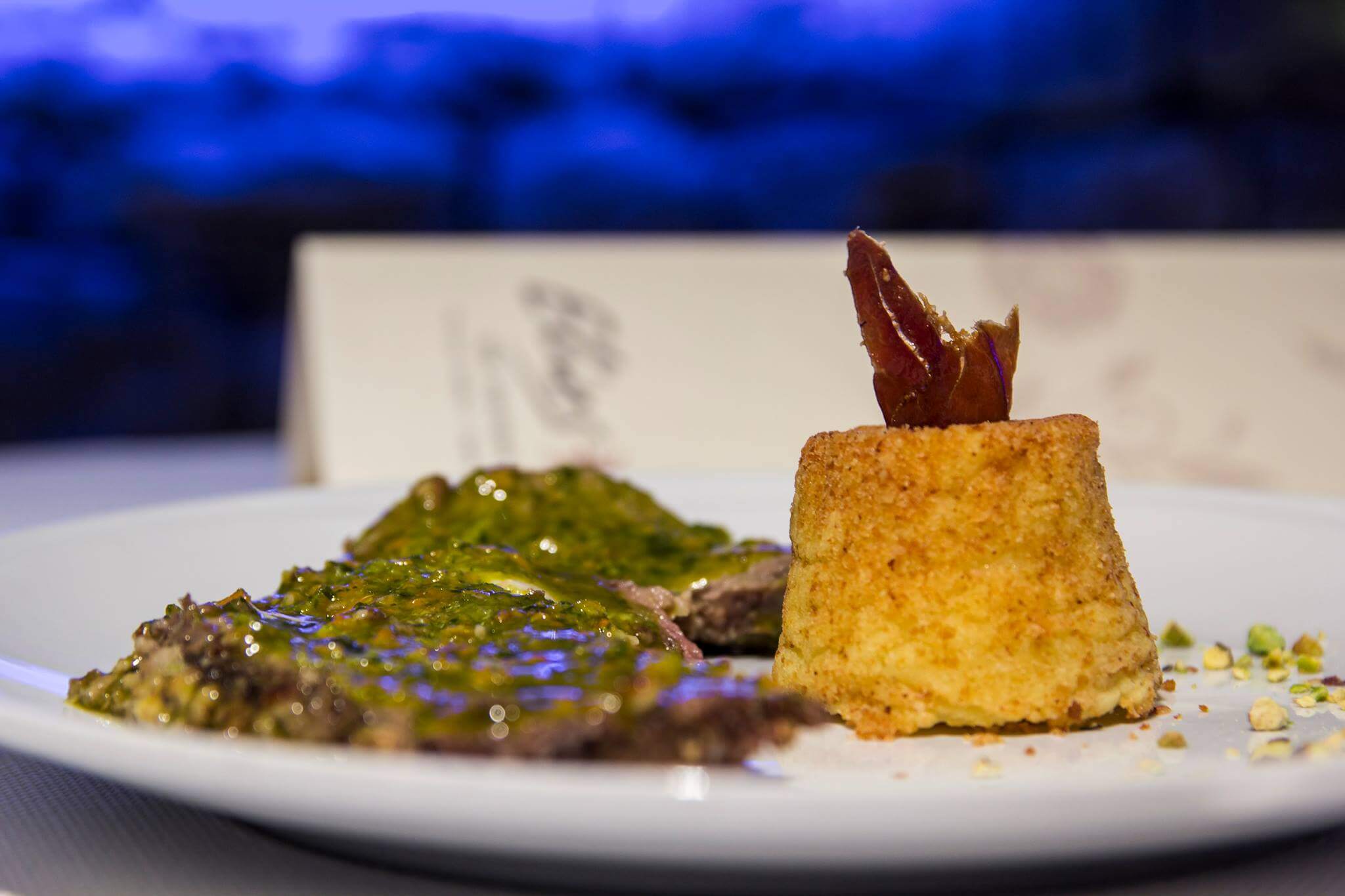 Un piatto con carne in salsa verde e tortino di patate con prosciutto in uno sfondo con luce blu