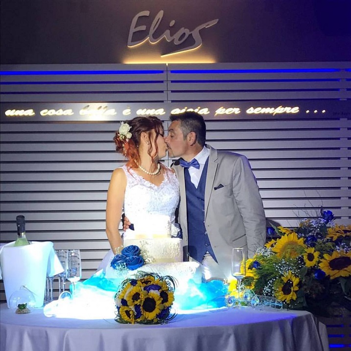 coppia di sposi celebra festa in vestiti eleganti davanti ad un tavolo con torta e fiori gialli e blu